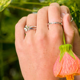 A person's hand wearing a silver and brass daisy ring and another silver flower band ring, holding a pink hanging flower.